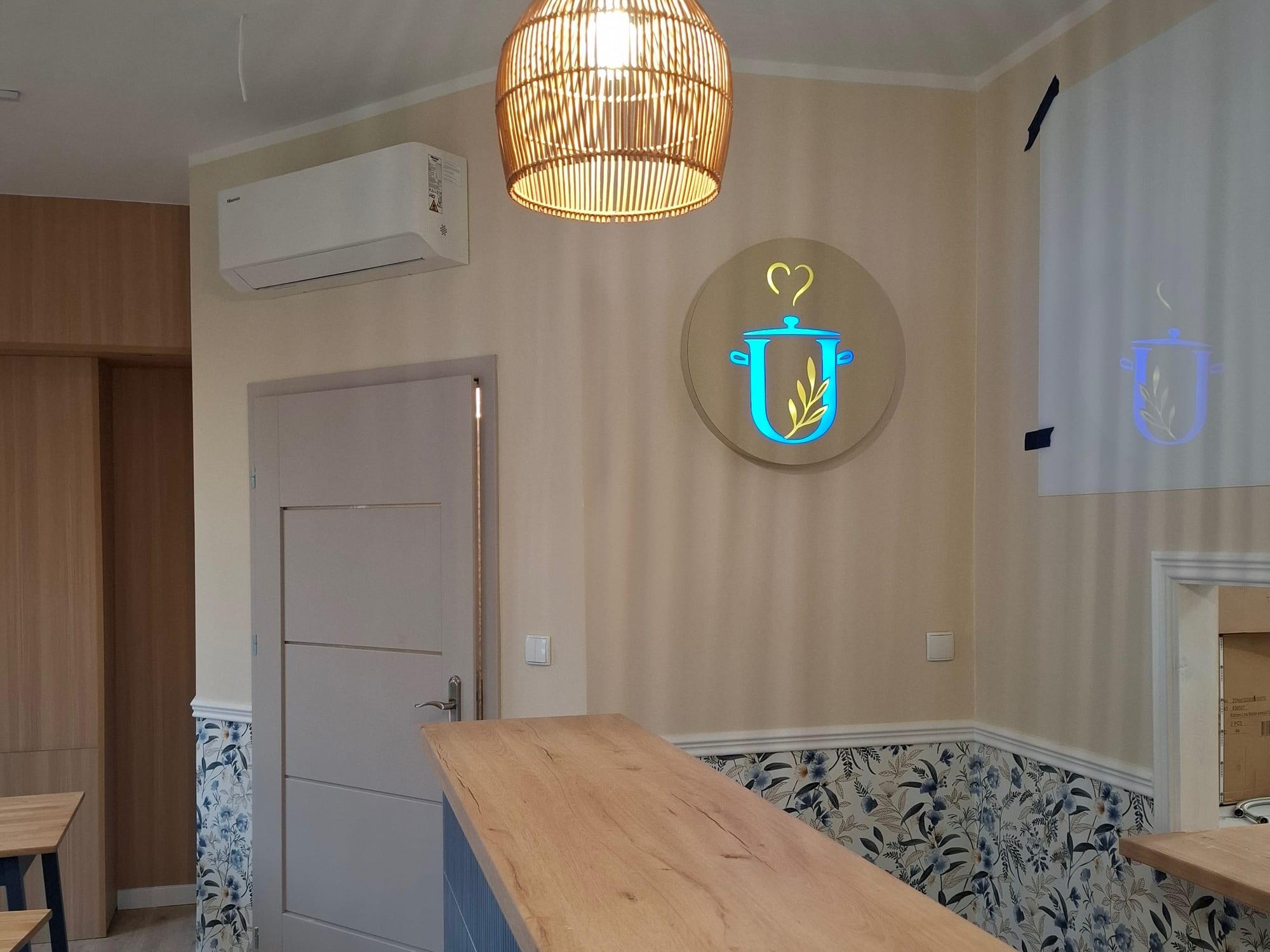 Okrągły, podświetlany szyld z logo uPICHcone – garnkiem z sercem – na ścianie sali bistro, nad barem wiklinowa lampa